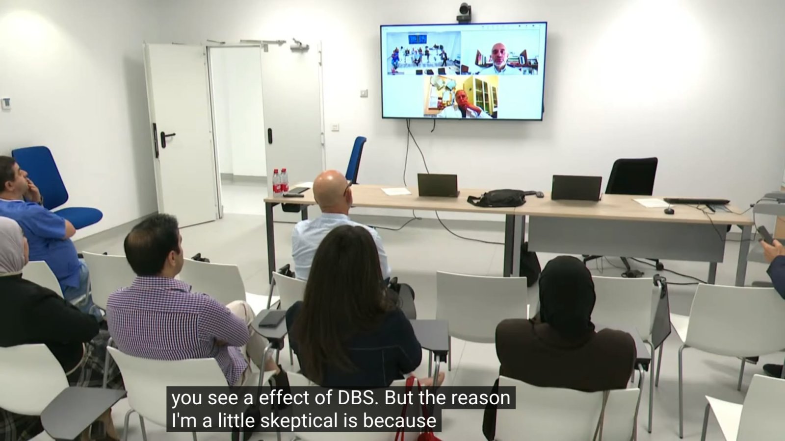 Personas sentadas de espaldas en una sala mirando una pantalla con una videollamada y subtítulos en inglés.