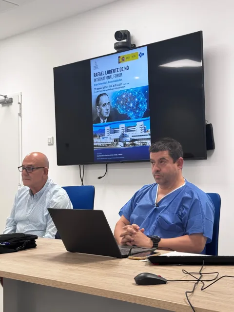 Dos hombres sentados ante una mesa con un portátil, uno con pijama sanitario azul, y tras ellos una pantalla con un cartel de un foro internacional.