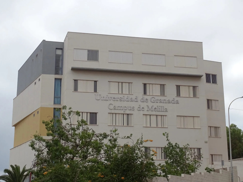 Edificio blanco de varias plantas con el rótulo Universidad de Granada Campus de Melilla y árboles delante.