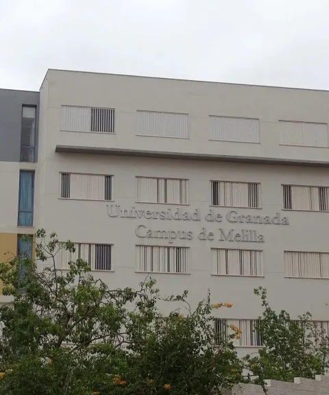 Fachada de un edificio blanco de varias plantas con ventanas y el rótulo Universidad de Granada Campus de Melilla, con arbustos delante.