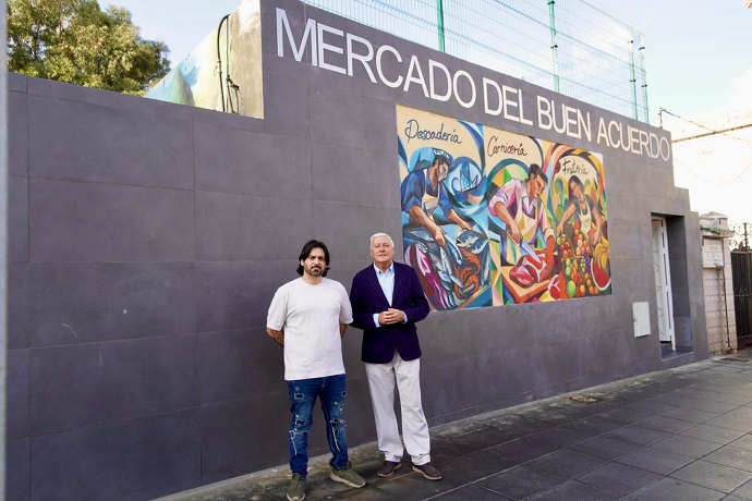 Dos hombres de pie ante una pared gris con el rótulo MERCADO DEL BUEN ACUERDO y un mural de pescadería, carnicería y frutería.
